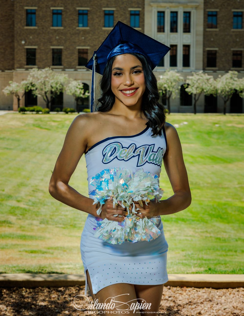 Young woman in graduation cap and cheerleader uniform smiling outdoors on campus green.
