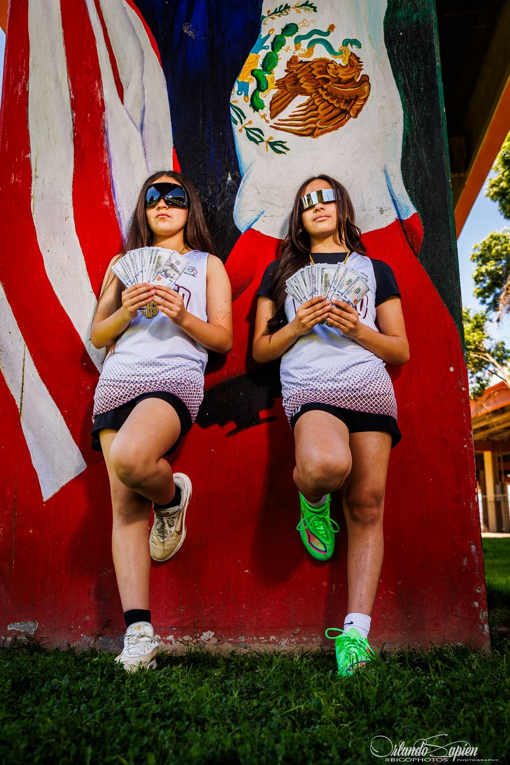 Two young women holding dollar bills pose against a colorful mural featuring the Mexican and American flags.