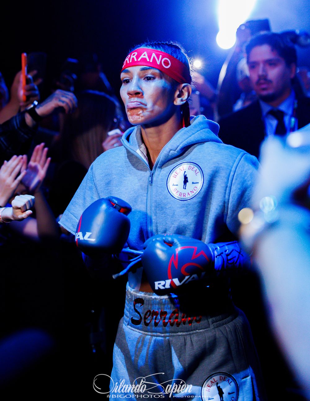 Boxer Serrano enters the ring wearing blue Rival gloves and a gray hoodie under dramatic stage lighting.
