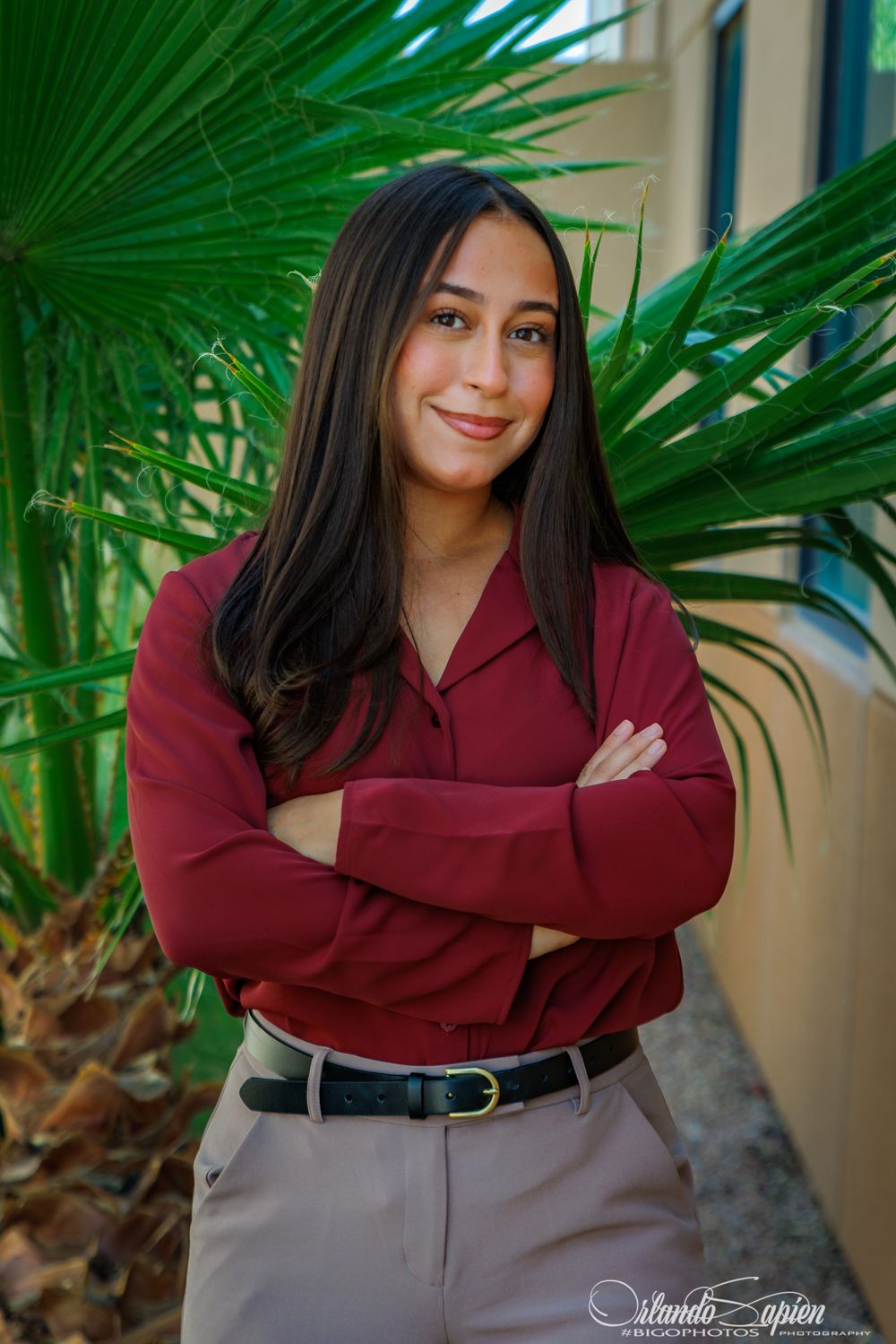 Young woman in maroon blouse with arms crossed smiling outdoors near tropical palm plants.