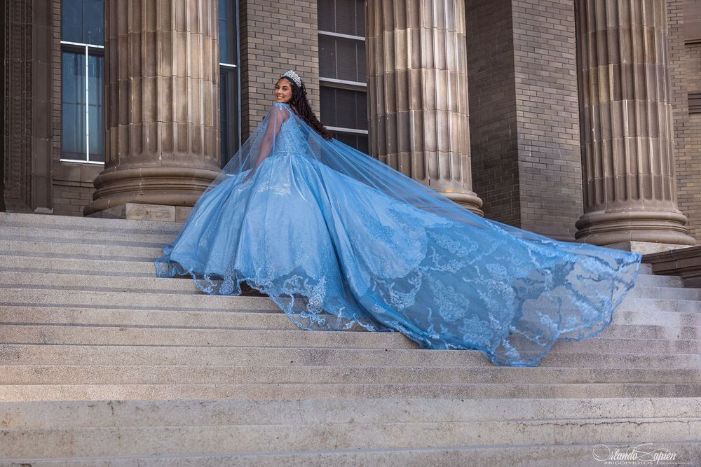 Woman in stunning blue ball gown with flowing train poses on grand stone steps before classical columns.