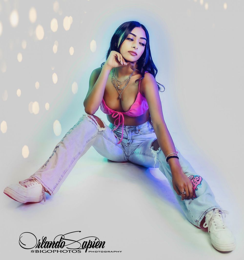 Person in white outfit and pink boots poses on floor with colorful bokeh lighting effects in studio setting.
