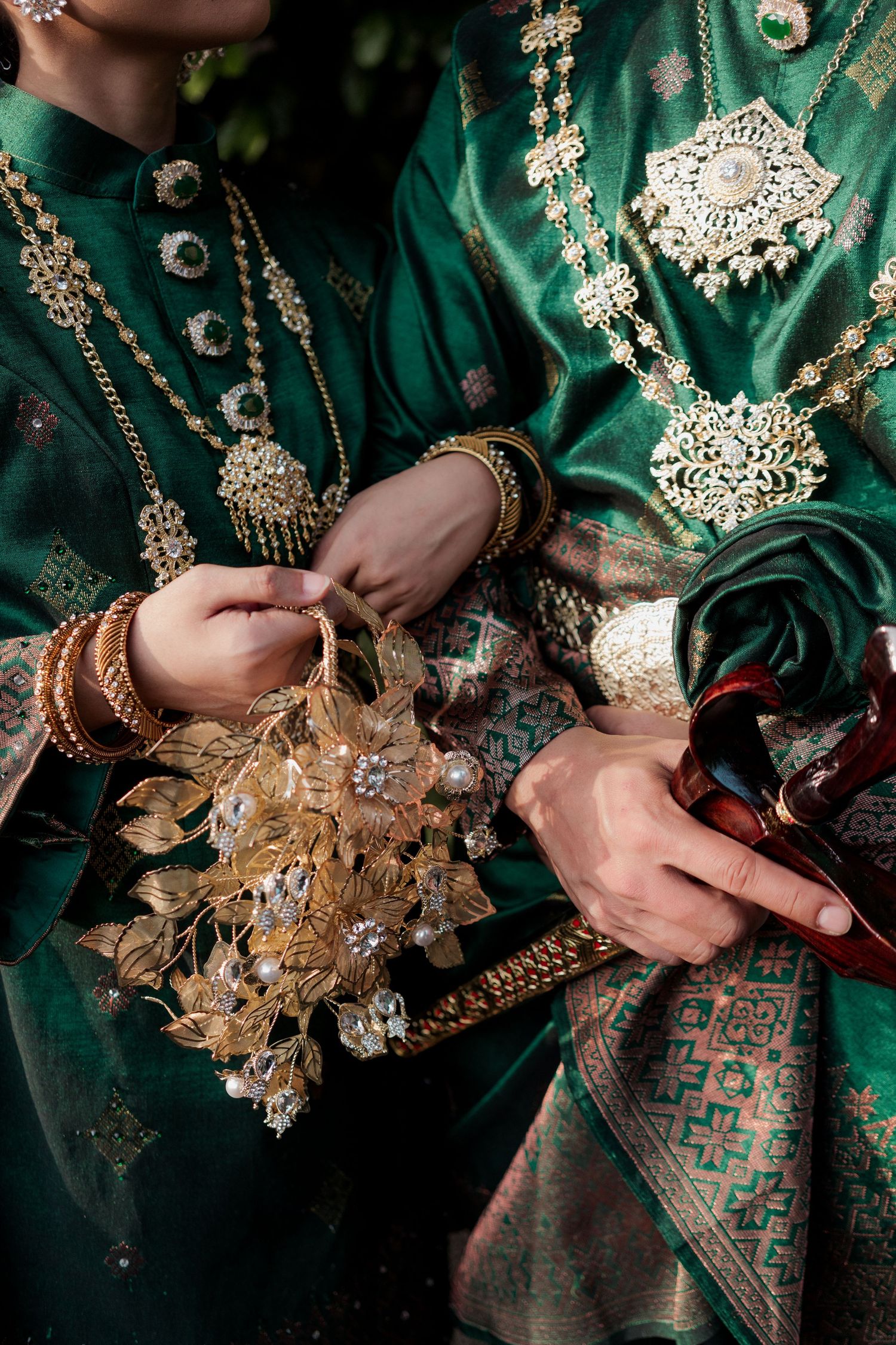 Two people in green traditional South Asian attire with gold jewelry holding an ornate floral gold bouquet.