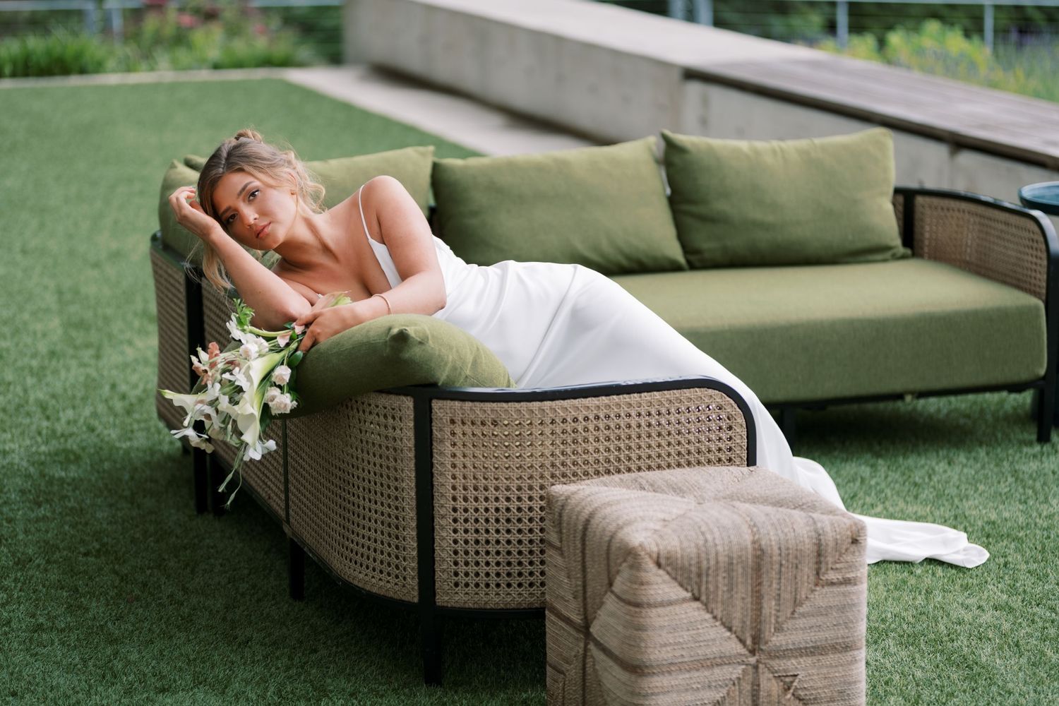 A person in a white dress rests on a modern curved rattan sofa with green cushions on artificial turf.