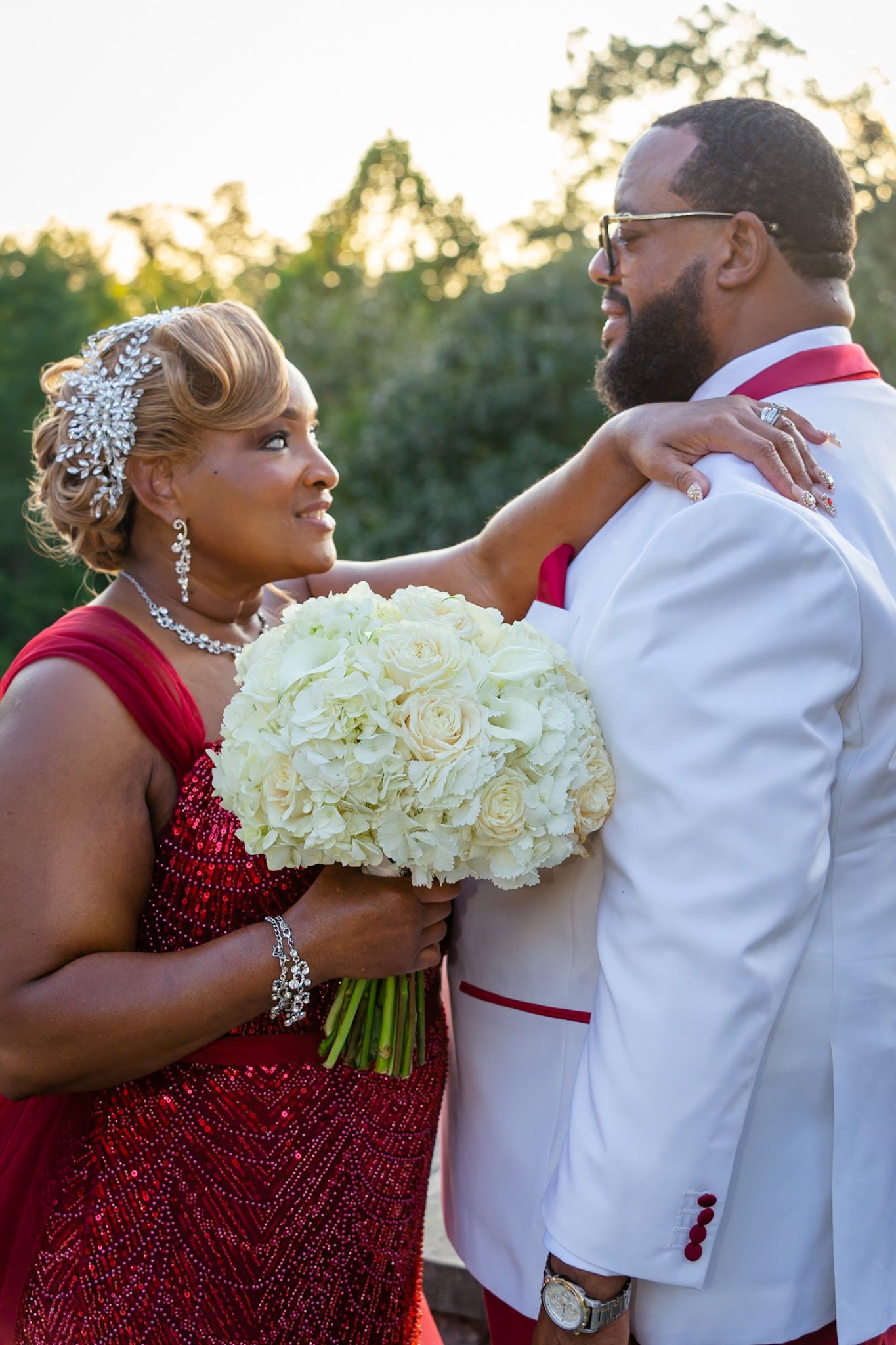 A wedding couple in red and white formal attire posing with a bouquet of white roses in an outdoor setting.