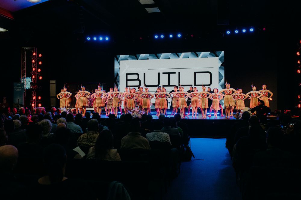 Performers in matching outfits on stage at a BUILD event with audience watching in a darkened venue.