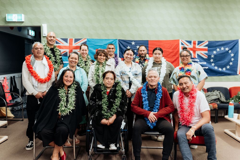 Group of people wearing leis seated and standing before New Zealand and Cook Islands flags.