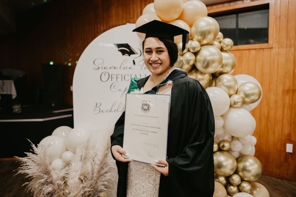 Smiling graduate in cap and gown proudly displaying diploma at University of Auckland ceremony.