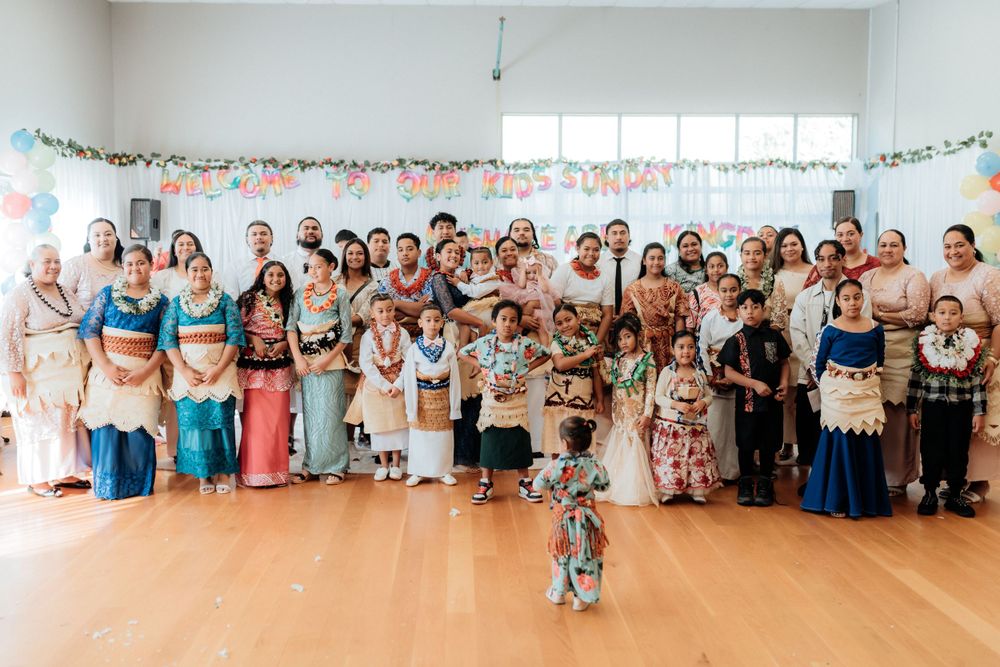Large family group photo in traditional Pacific Island attire at a decorated community hall celebration.