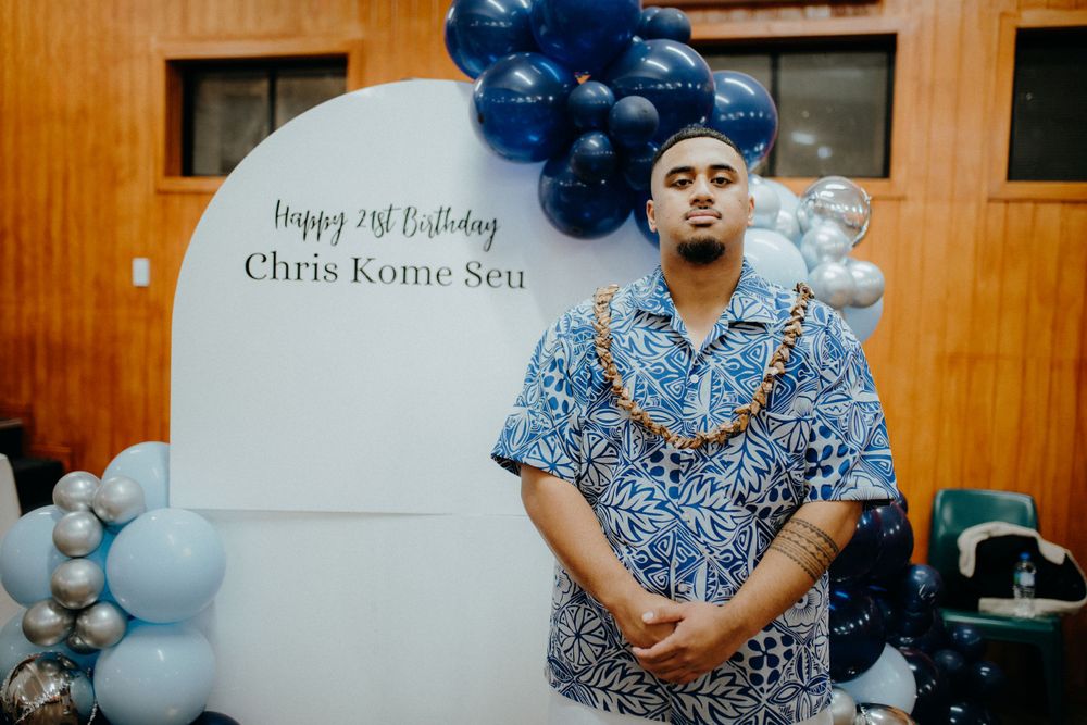 Man in blue Hawaiian shirt stands before Happy 21st Birthday Chris Kome Seu balloon backdrop.