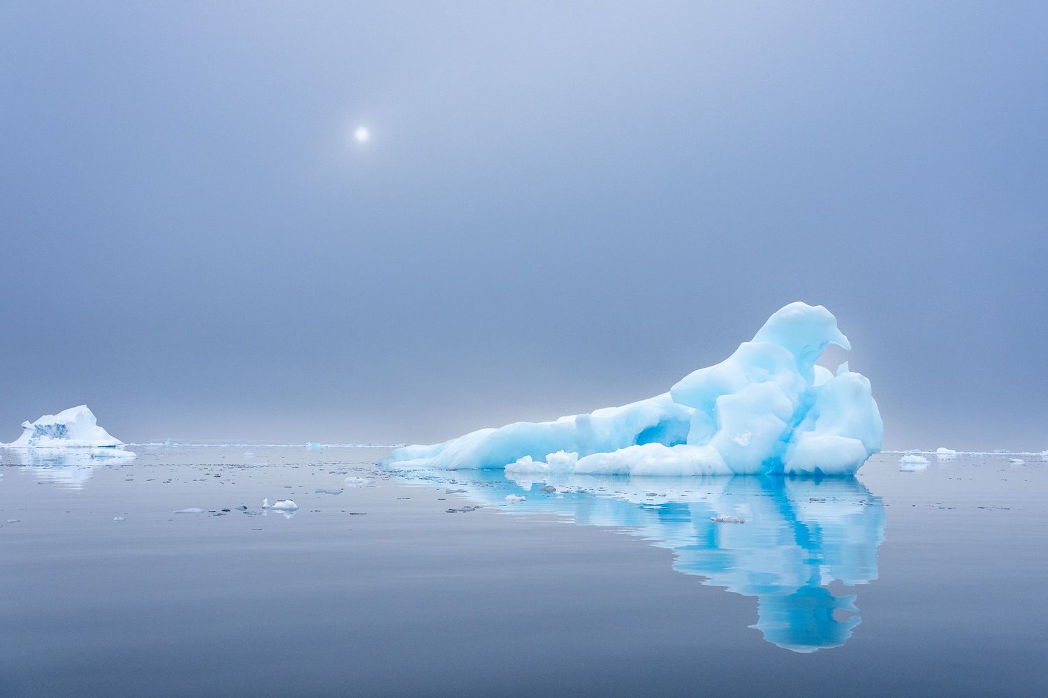 A vivid blue iceberg reflects on calm Arctic waters beneath a misty, pale sky with a faint sun glowing above.