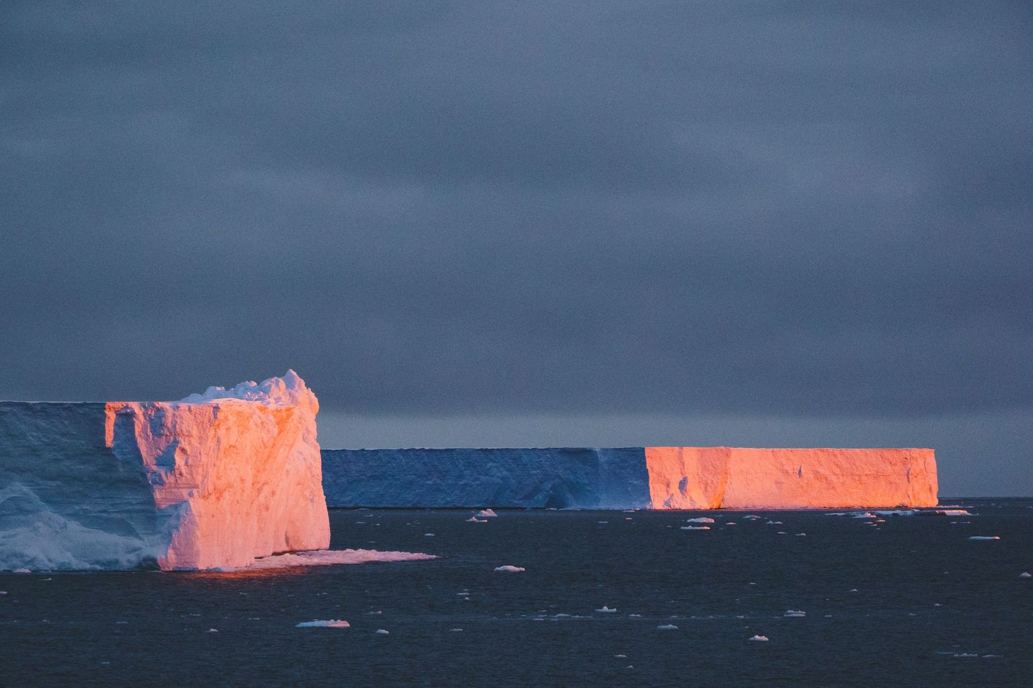 Massive flat-topped icebergs glowing orange in sunset light against a dramatic stormy sky in Antarctica.