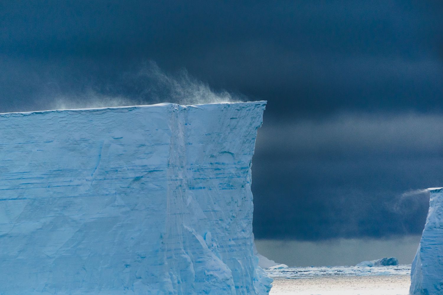 Massive blue iceberg with snow blowing off its flat top against a dramatic stormy dark sky in Antarctica.