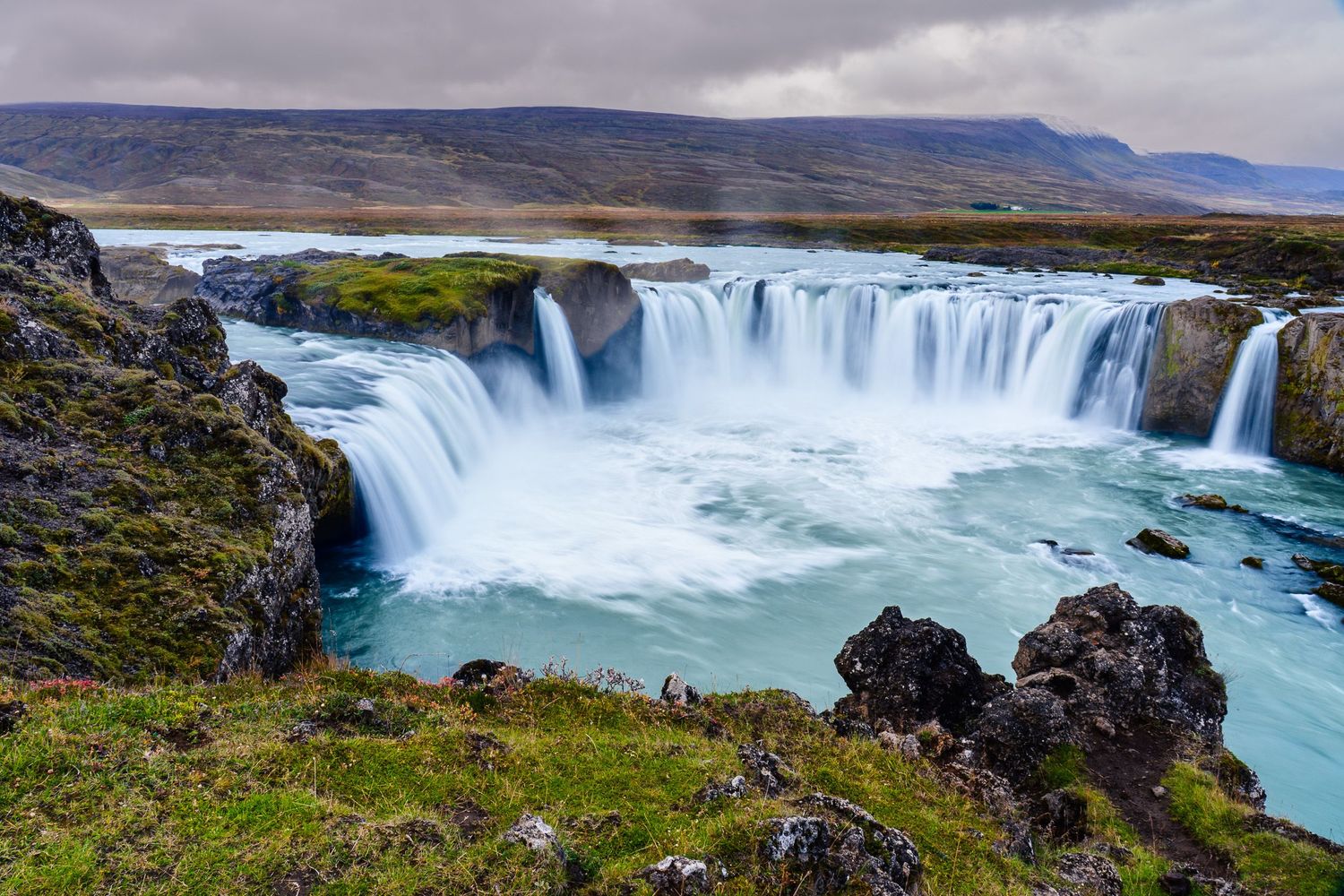 Majestic Godafoss waterfall in Iceland cascading into a turquoise river surrounded by rocky terrain under cloudy skies.