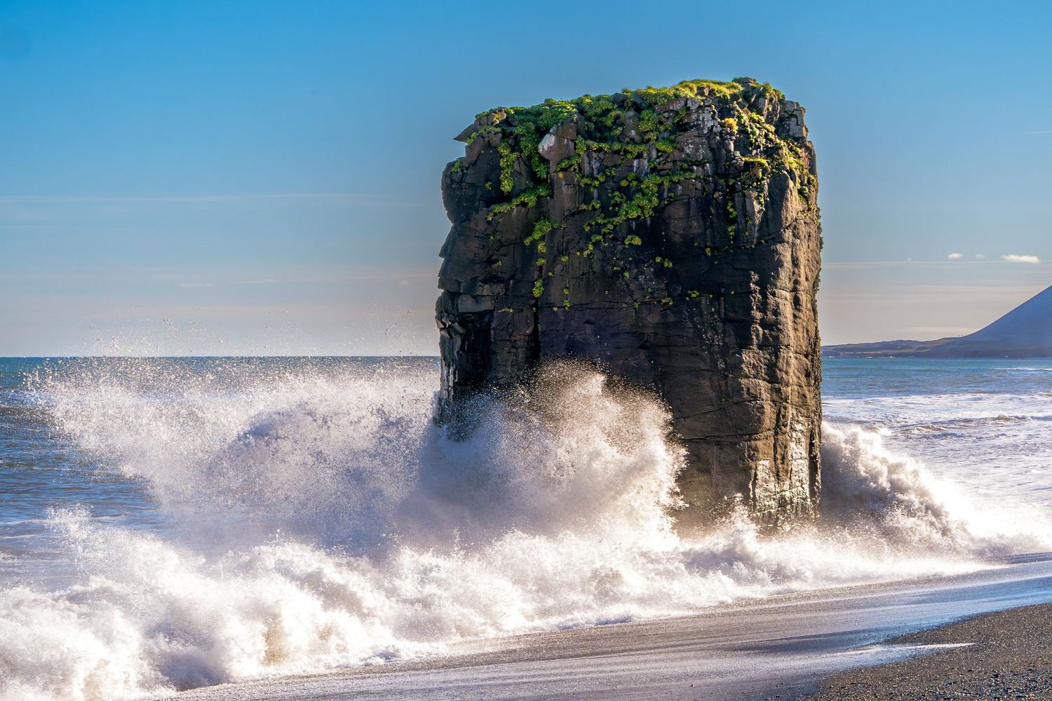 A dramatic sea stack rock formation topped with green vegetation being crashed by powerful ocean waves on a black sand beach.