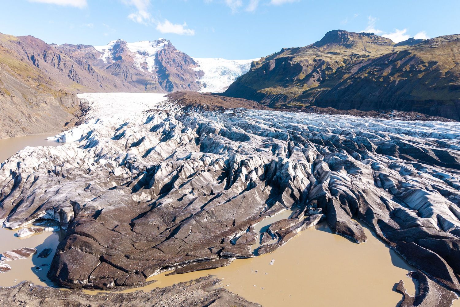 Aerial view of a vast glacier with dark volcanic ash streaks flowing into a glacial lagoon surrounded by mountains in Iceland.