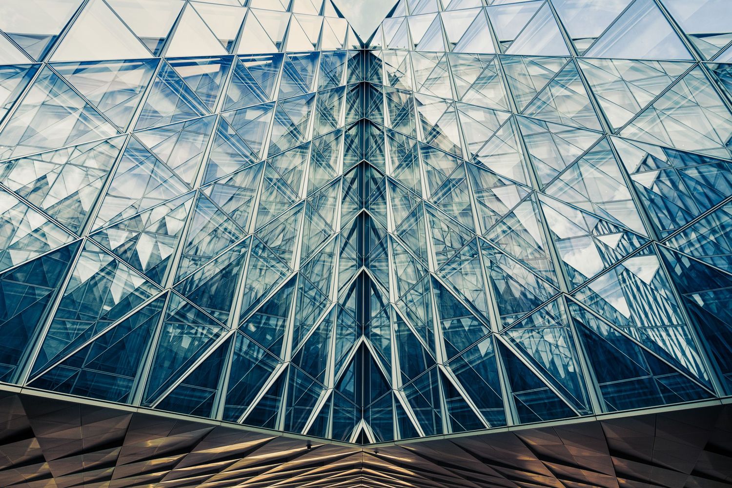 Abstract geometric glass building facade with triangular steel framework reflecting blue sky in modern architecture.
