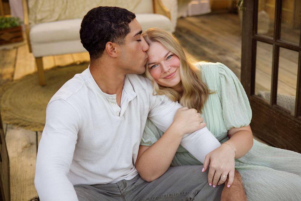 A young interracial couple sitting together, man kissing woman on cheek, wearing casual pastel outfits indoors.
