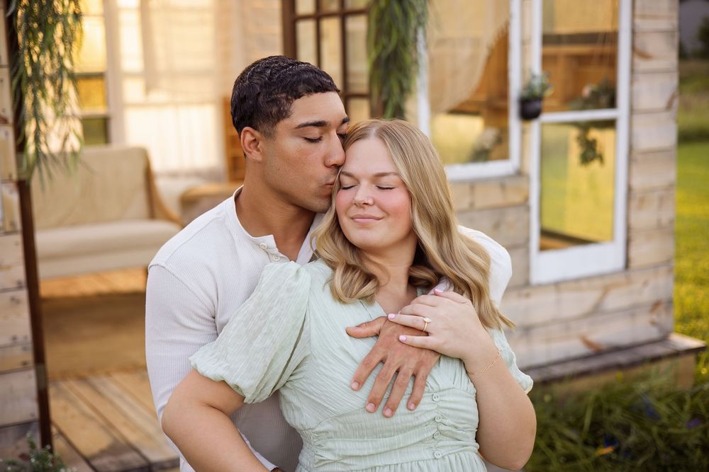 A young couple shares a tender moment outdoors, man kissing womans cheek while embracing her near a house.