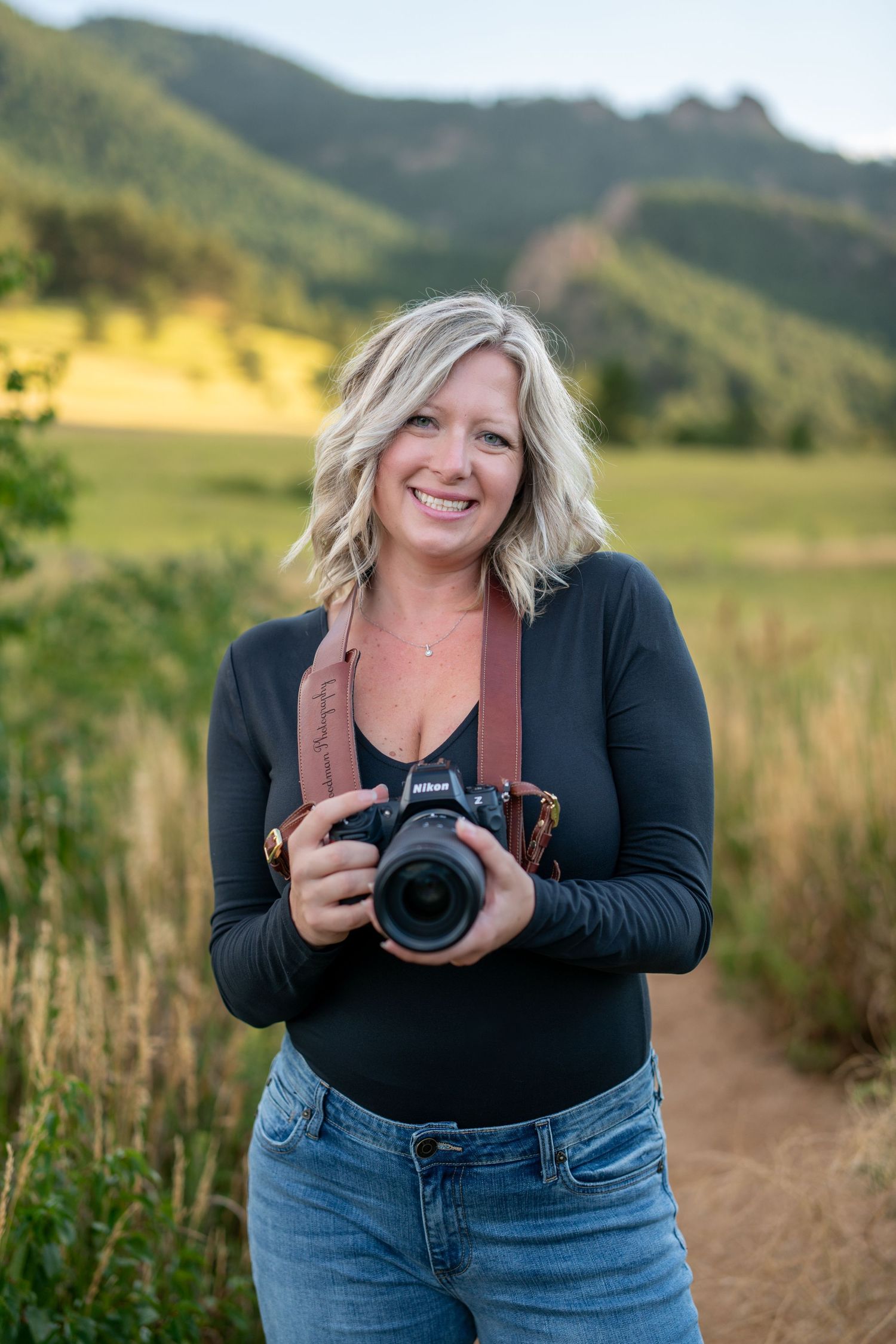 Photographer with camera standing on trail with mountain landscape in background.