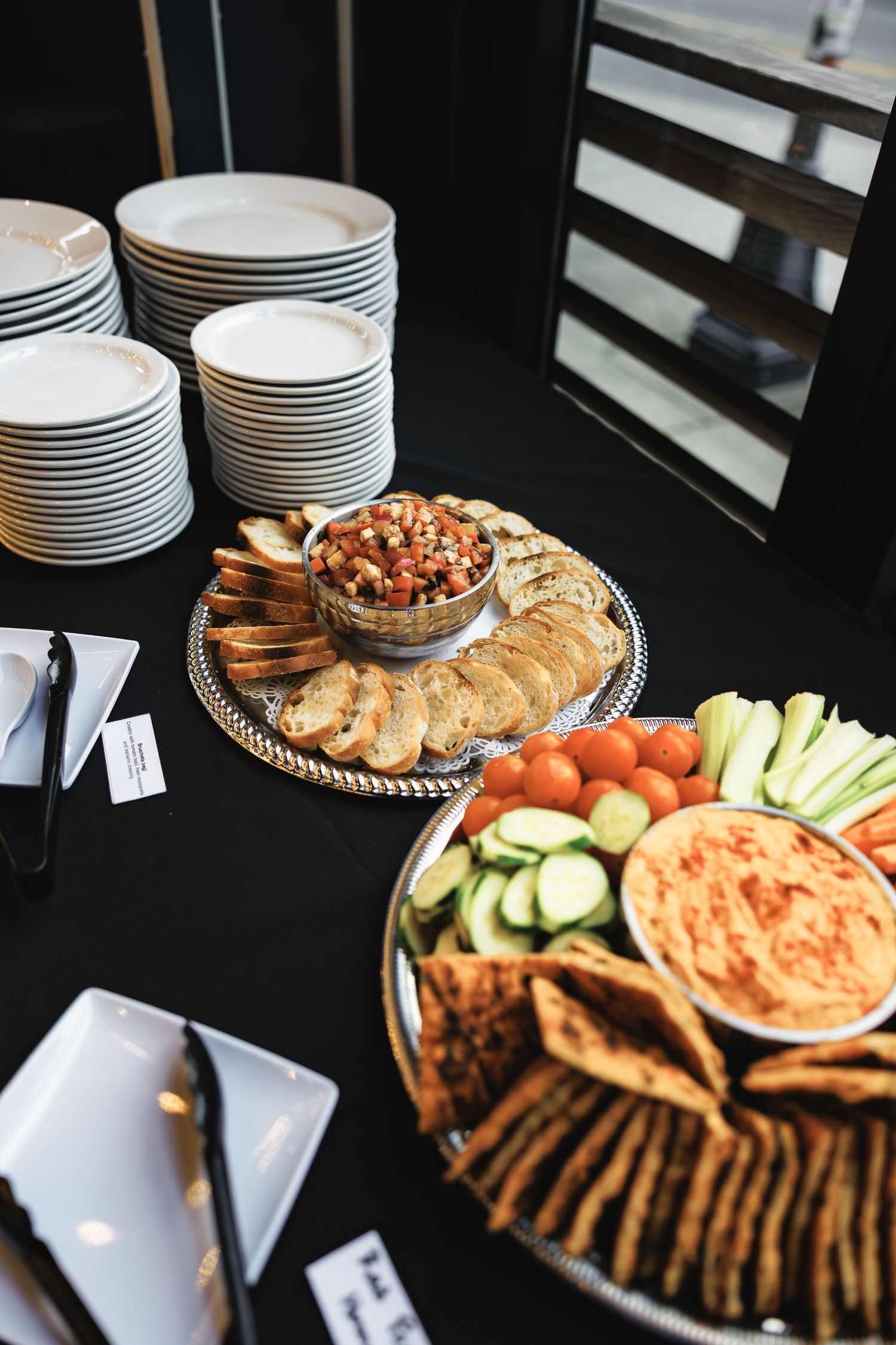 Appetizer platters with crackers, vegetables, and dips served alongside stacked white plates.