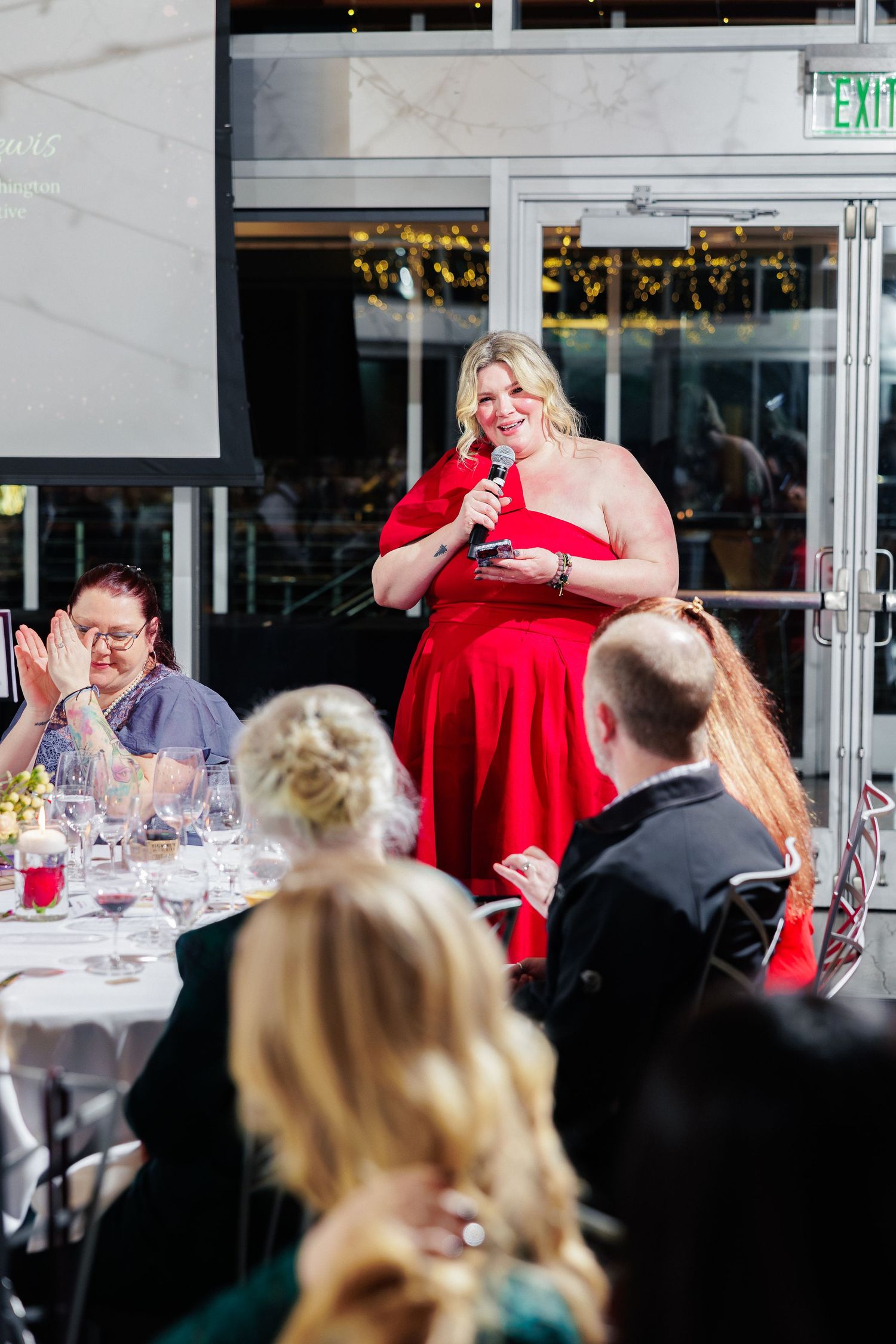 A woman in a red dress delivers a speech at an elegant dinner event with guests seated at decorated tables.