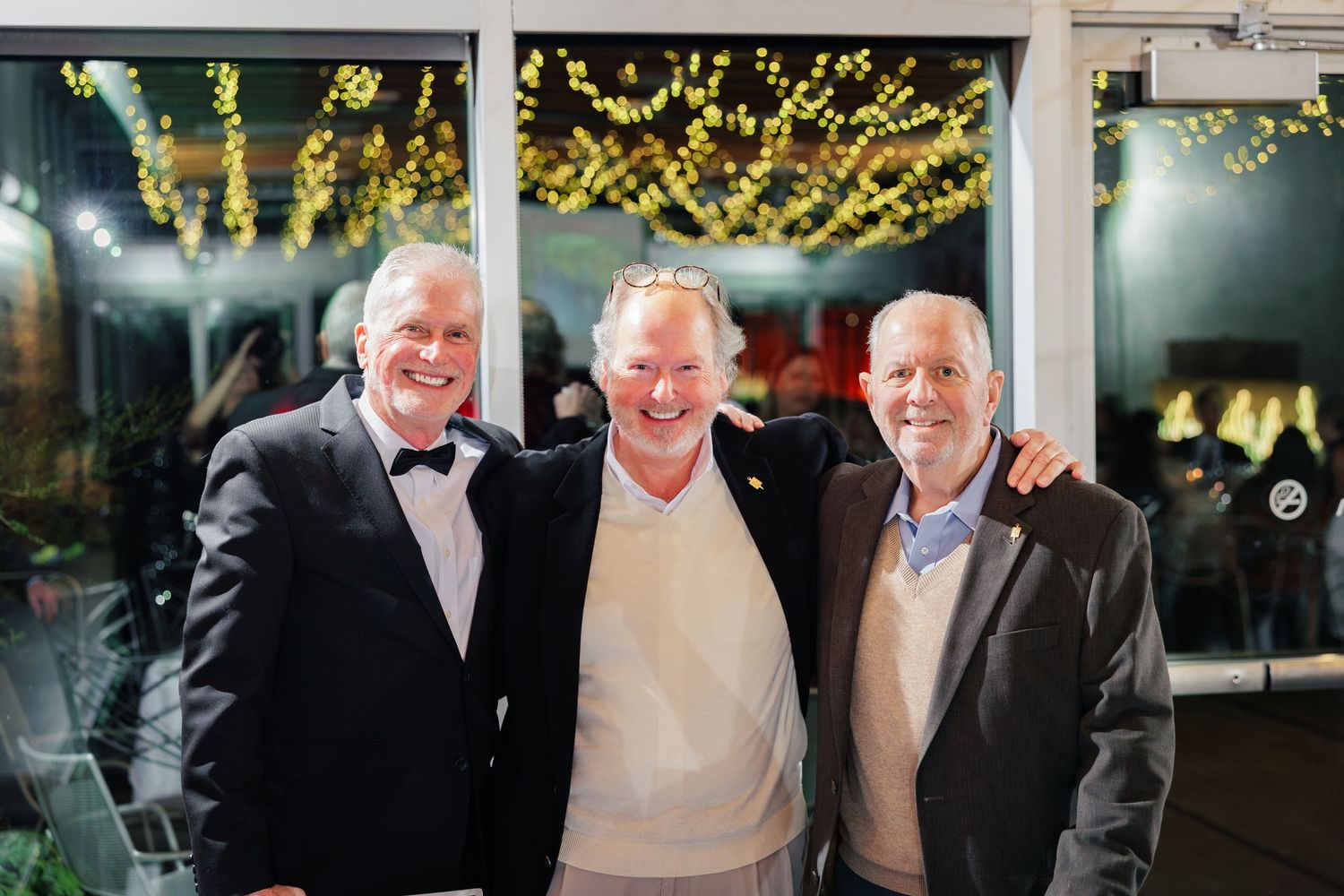 Three men in formal attire stand together smiling at an evening event with decorative lights visible behind them.