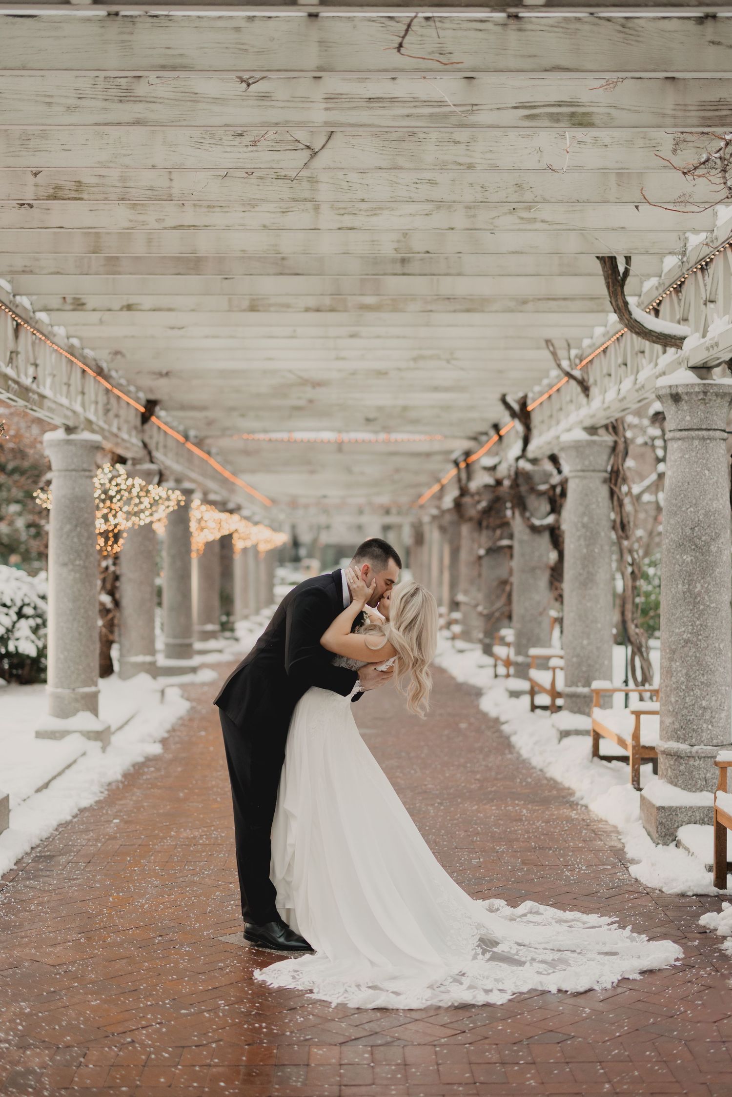 A couple walks hand in hand along a path beside a grand Mediterranean-style villa with mountain views.