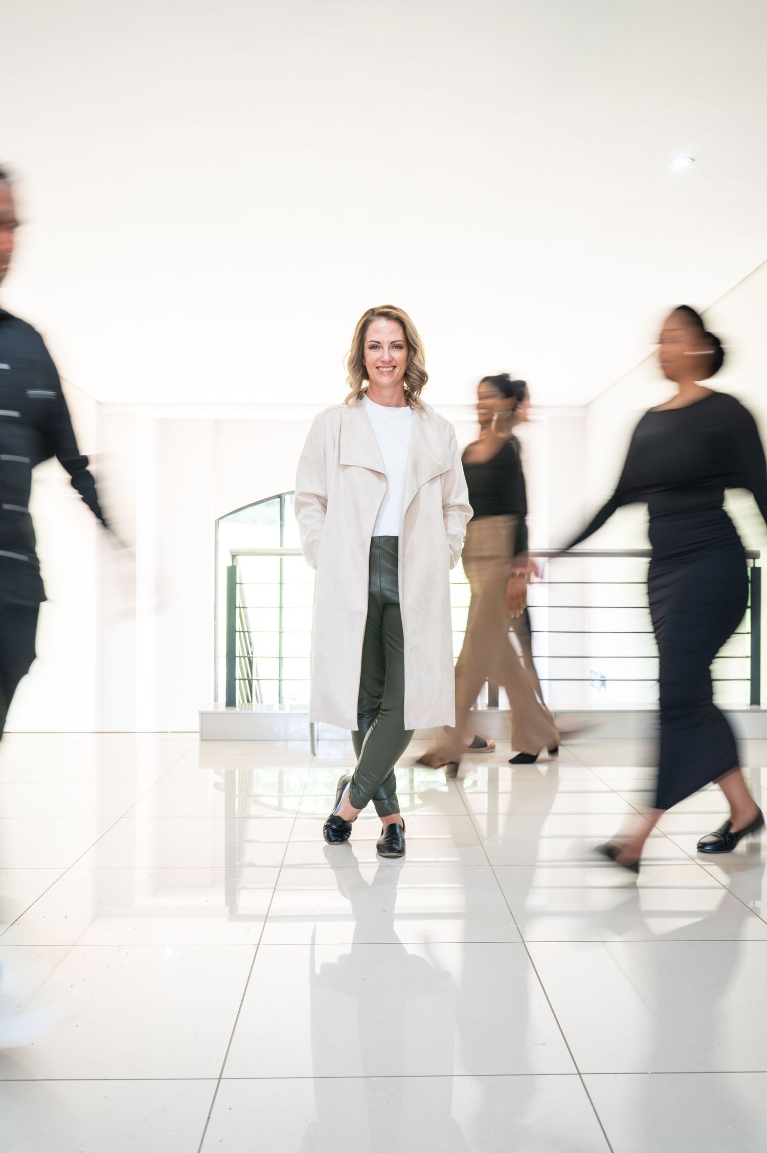 Confident businesswoman during her personal branding photoshoot standing still amid blurred colleagues walking in modern office lobby.