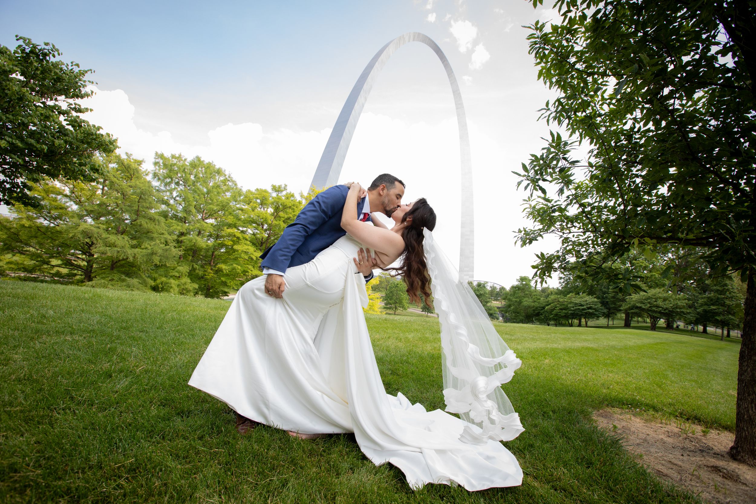 Dramatic wedding photo with couple kissing under the St. Louis Arch in lush green garden.