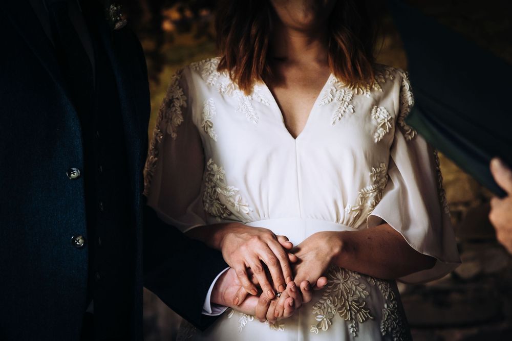 A bride in a white lace dress holds her hands together while standing in soft, moody lighting.