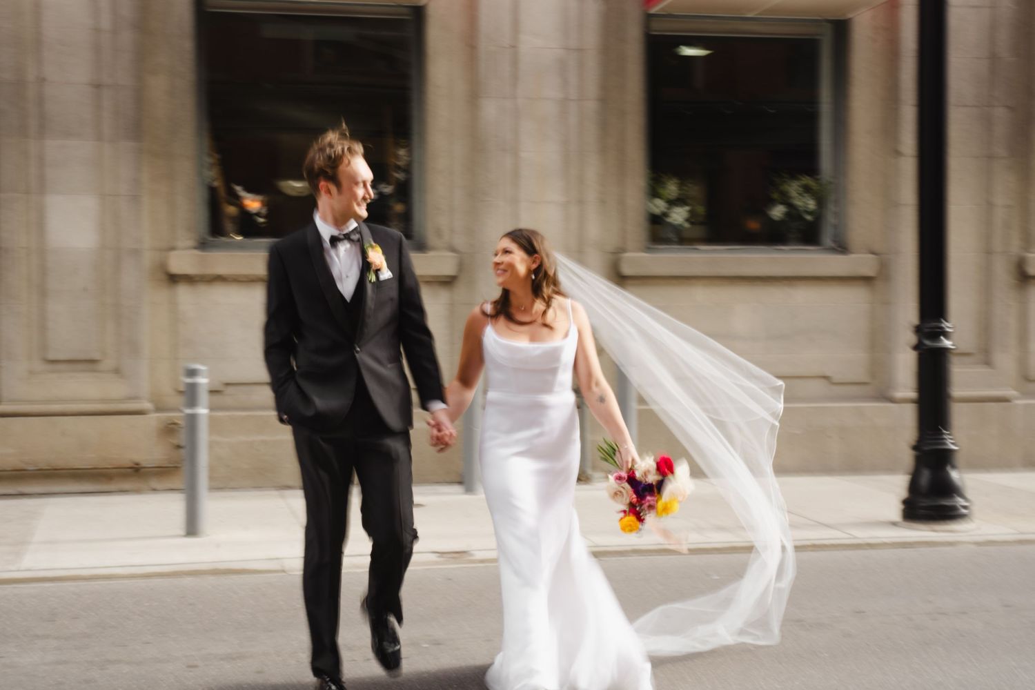 Wedding couple walking and posing outside classical stone building with flowing veil.