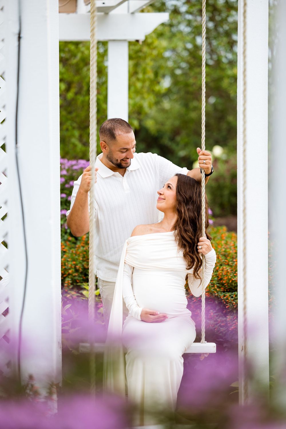 Pregnant woman in white dress sits on garden swing while partner stands beside her among colorful flowers.