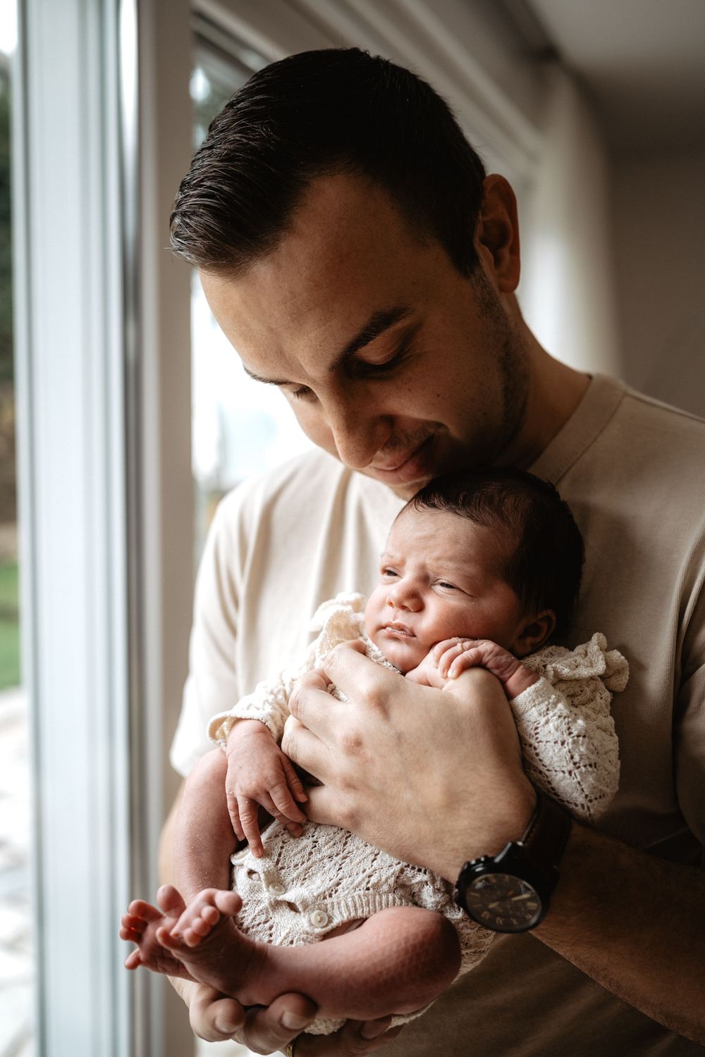 A father tenderly kisses his newborn baby in a series of intimate portraits by a window.