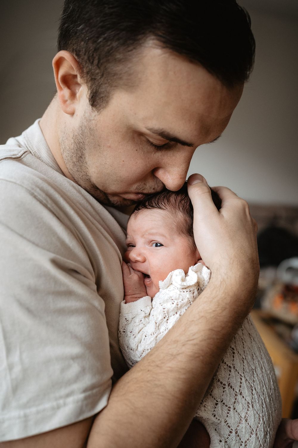 A tender moment shows a parent in a beige shirt holding and kissing a newborn baby swaddled in white lace.