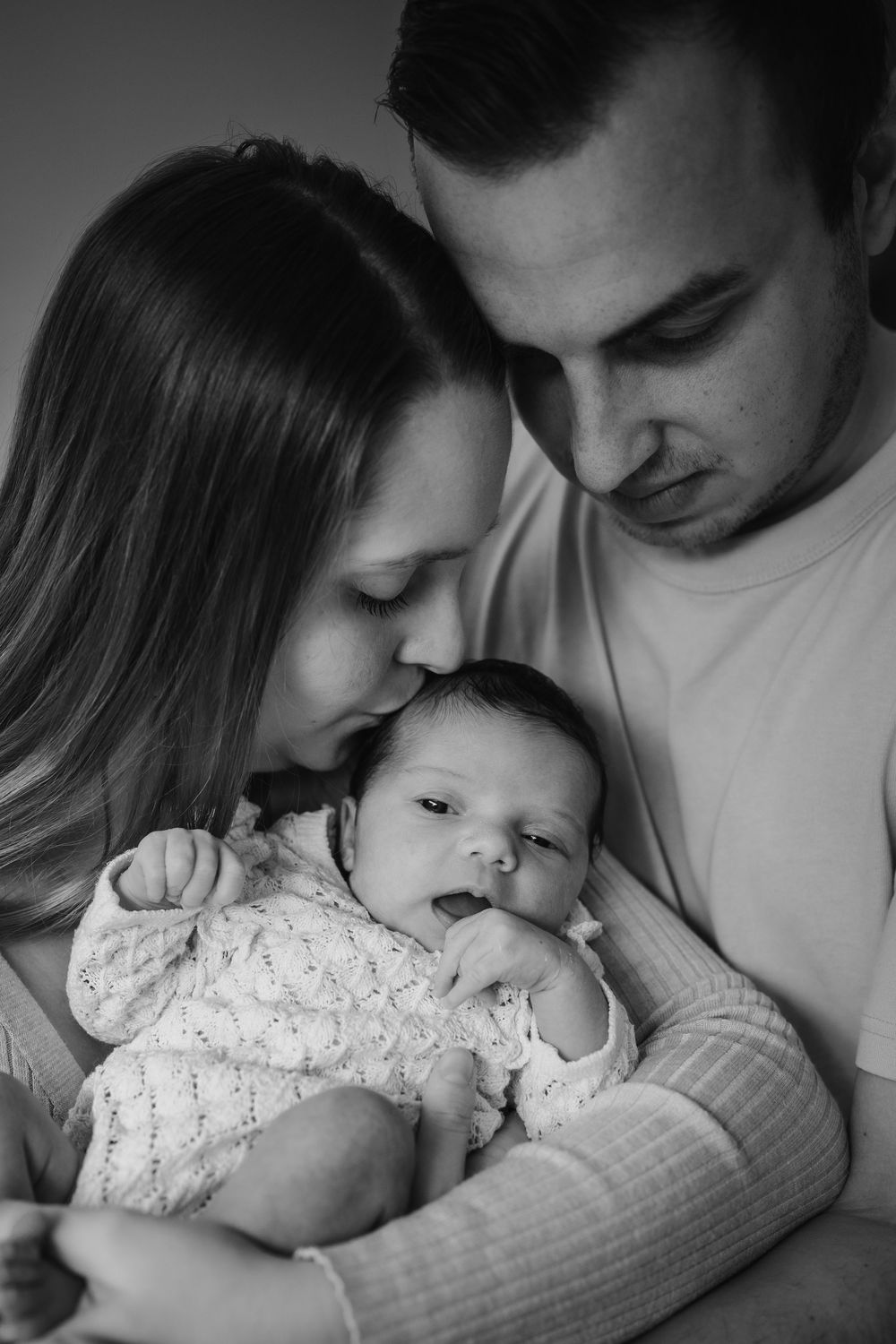 Black and white intimate portrait of parents holding and gazing lovingly at their newborn baby in a lace outfit.