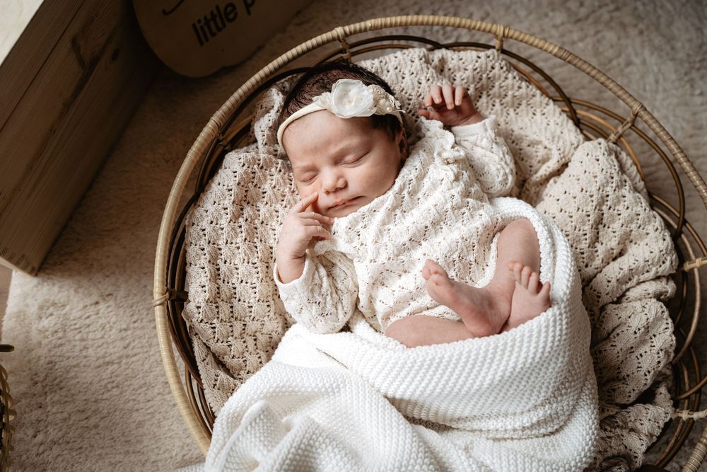 A sleeping newborn baby swaddled in white lace blankets rests peacefully in a rustic wooden basket.