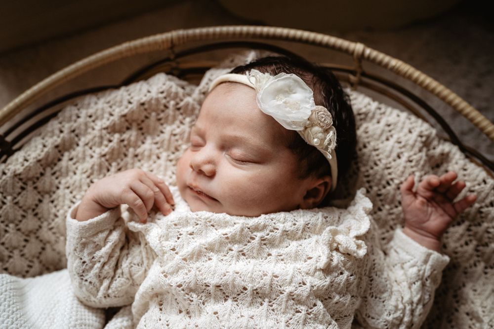 Sleeping baby wearing cream knit sweater and white bow headband rests peacefully in woven basket on textured blanket.