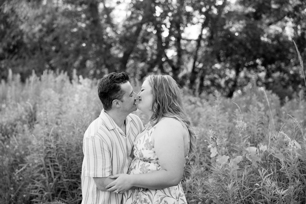 A romantic couple embraces in a field of tall grass and wildflowers during a black and white outdoor photo session.