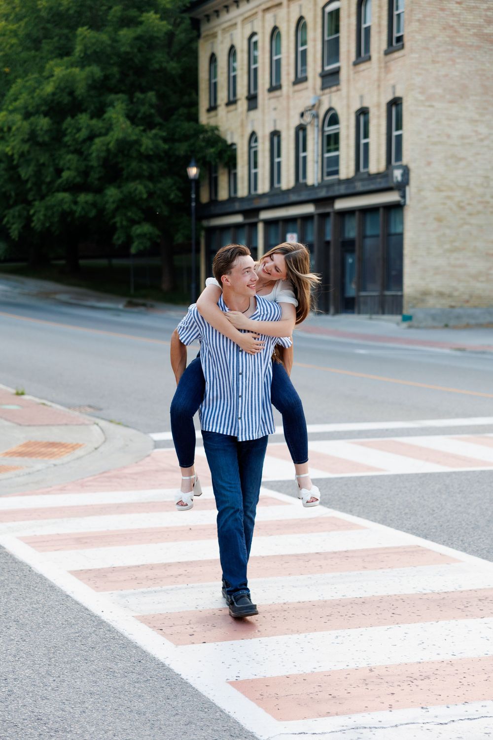 A couple shares a playful piggyback ride while crossing a pink and white striped crosswalk in front of a historic building.