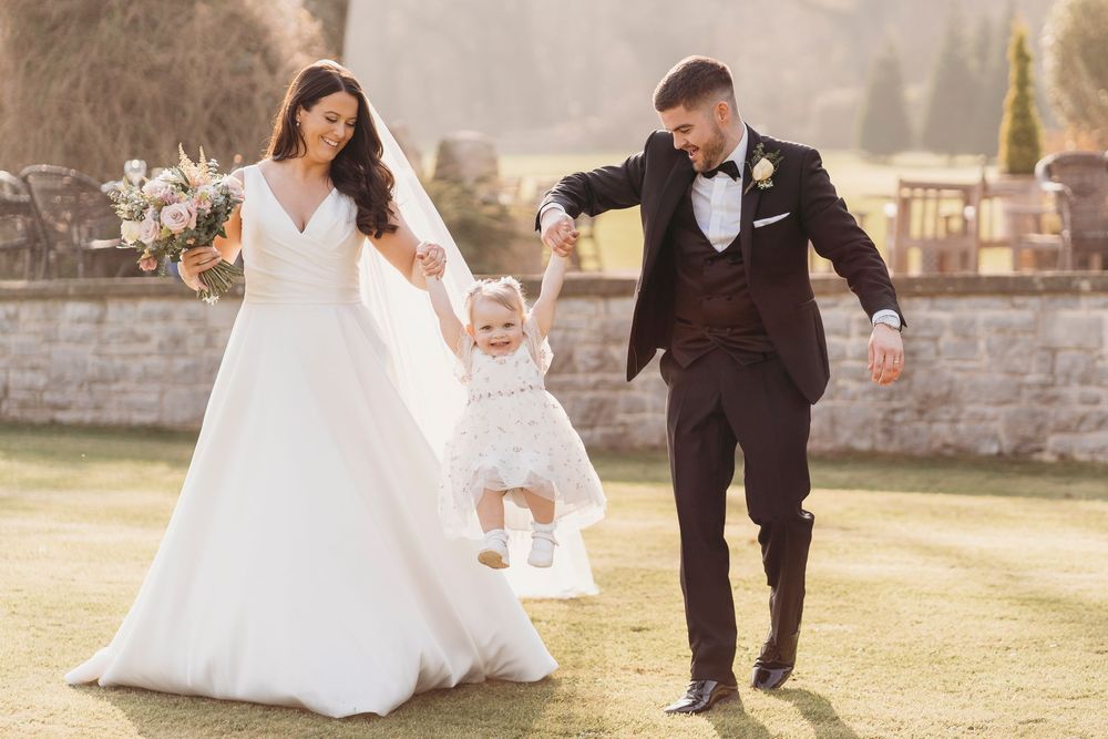 Bride, groom and their young daughter walking across a lawn at golden hour.