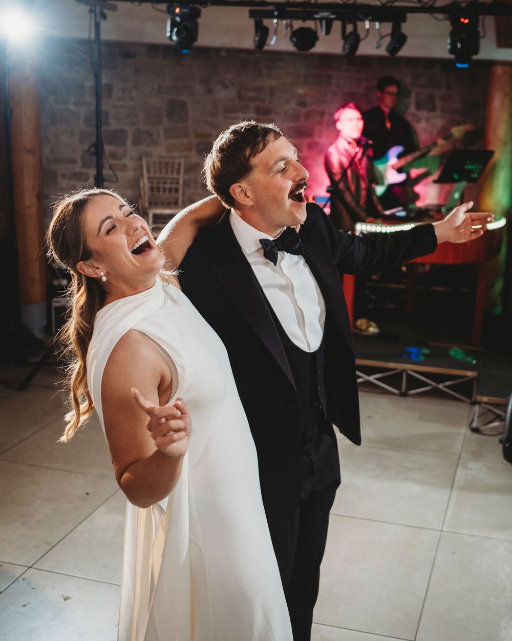 Newlyweds dancing on the dance-floor with their arms out and singing along with the band.