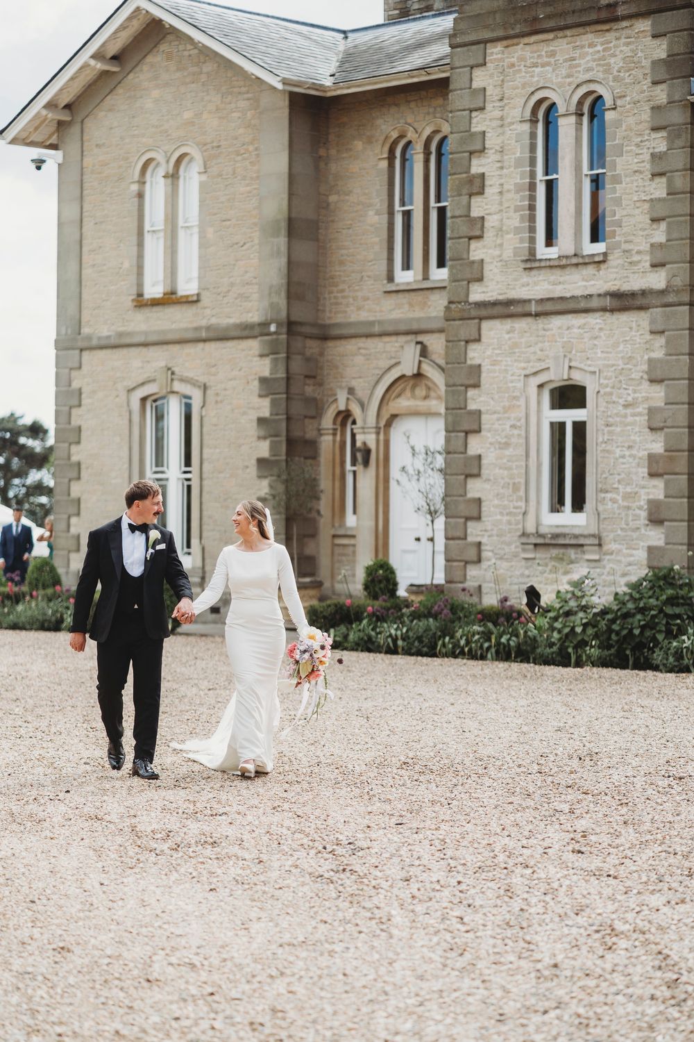 A bride and groom walk hand-in-hand across a gravel driveway in front of the historic, stone-built St Tewdrics House in Chepstow. The bride