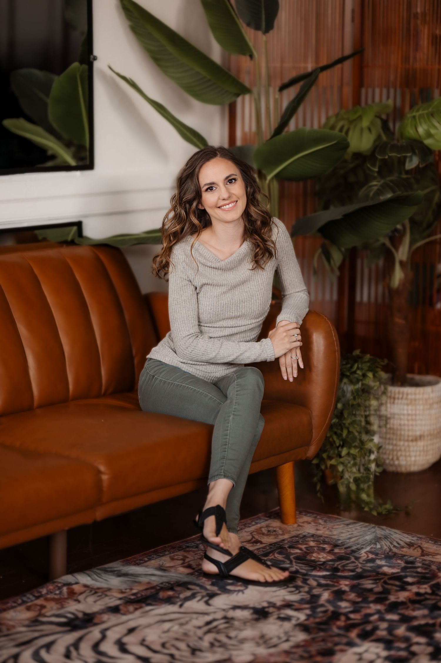Portrait of woman in gray shirt against sitting on a brown leather couch, smiling at camera with one leg crossed over the other.
