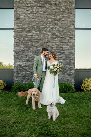 A couple poses with their two dogs against a modern stone wall on their wedding day.