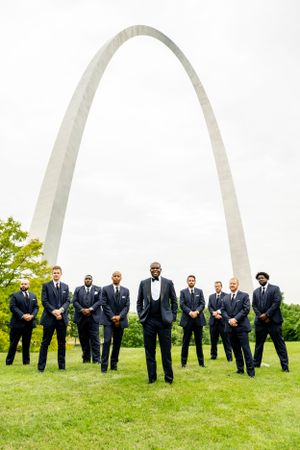 Wedding party in navy suits poses on a grassy hill with the iconic St. Louis Arch in the background.