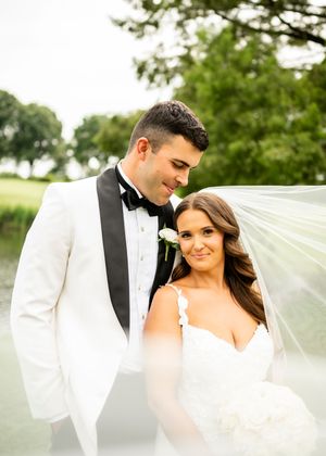 Romantic wedding portrait with flowing veil against green outdoor backdrop.