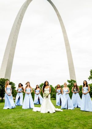Bridesmaids in light blue dresses pose together on a green lawn with the St. Louis Arch visible.
