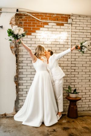 Couple in white wedding attire dances and celebrates against rustic brick wall backdrop.