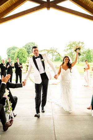 Wedding party performing choreographed dance moves under wooden beamed pavilion.